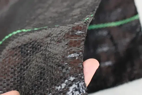 Hand holding the edge of dark woven geotextile fabric with a green marker thread.